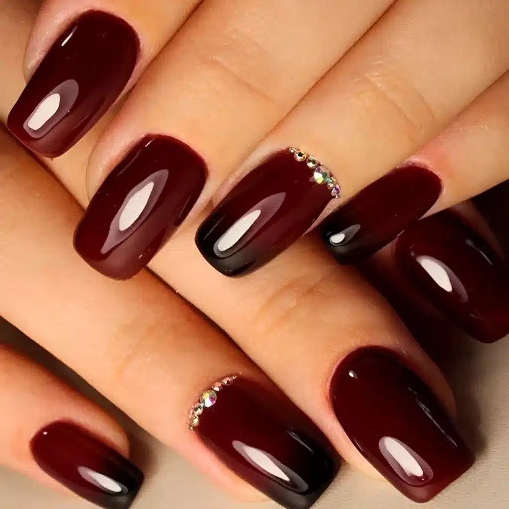 Hands displaying glossy burgundy nails with gemstones on both ring fingers