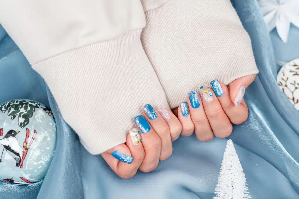 Hands with winter-themed nail art featuring blue and silver polish, snowflake designs, and small gem accents. Hands with winter-themed nail art featuring blue and silver polish, snowflake designs, and small gem accents.